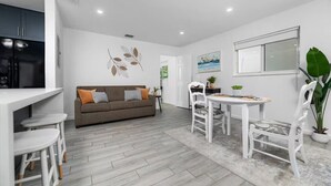 A modern kitchen with white cabinetry, a grey sofa with orange cushions, a dining table with white chairs, and a large green plant. - Your Downtown Getaway 2 (Fort Lauderdale)