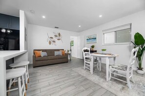 Eine moderne Küche mit weißen Schränken, ein graues Sofa mit orangefarbenen Kissen, ein Esstisch mit weißen Stühlen und eine große, grüne Pflanze.