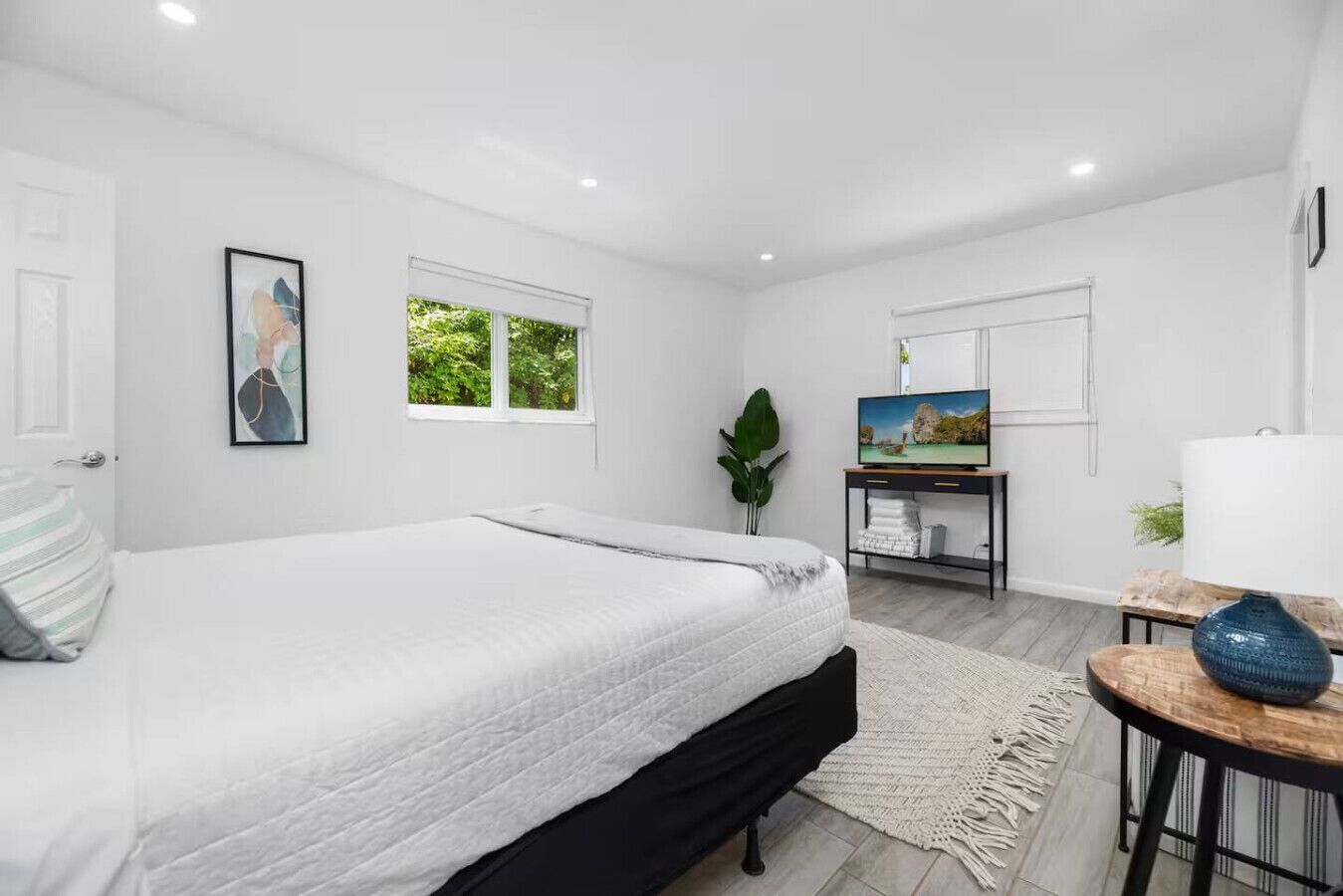 A modern bedroom with a large bed, a TV, a small table, and a window with a view of greenery.