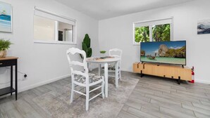 A room with a dining table and chairs, a TV on a wooden stand, and a window with a view of greenery. - Your Downtown Getaway 2 (Fort Lauderdale)