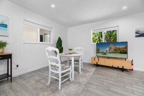 Ein Zimmer mit Esstisch und Stühlen, einem Fernseher auf einem Holzboden und einem Fenster mit Blick auf Grünflächen.