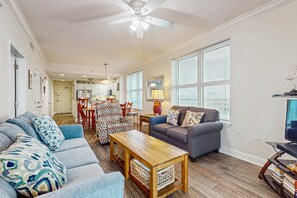 A living room with a sofa, a coffee table, a TV, and a dining area with chairs and a table. - Corner Gulf/beachfront condo with pool, hot tub, sauna, & private balcony (Gulf Shores)