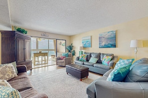 A living room with a dark brown sofa, a grey sofa, a wooden cabinet, and a dining area with a table and chairs. - Beachfront condo with enclosed balcony, pool, views, central AC, & grill (Gulf Shores)