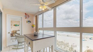 Une pièce dotée d’une grande fenêtre donnant sur la plage, d’un ventilateur de plafond et d’une petite table avec une chaise.