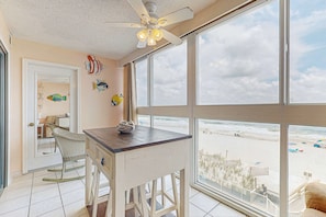 A room with a large window overlooking the beach, a ceiling fan, and a small table with a chair. - Beachfront condo with enclosed balcony, pool, views, central AC, & grill (Gulf Shores)