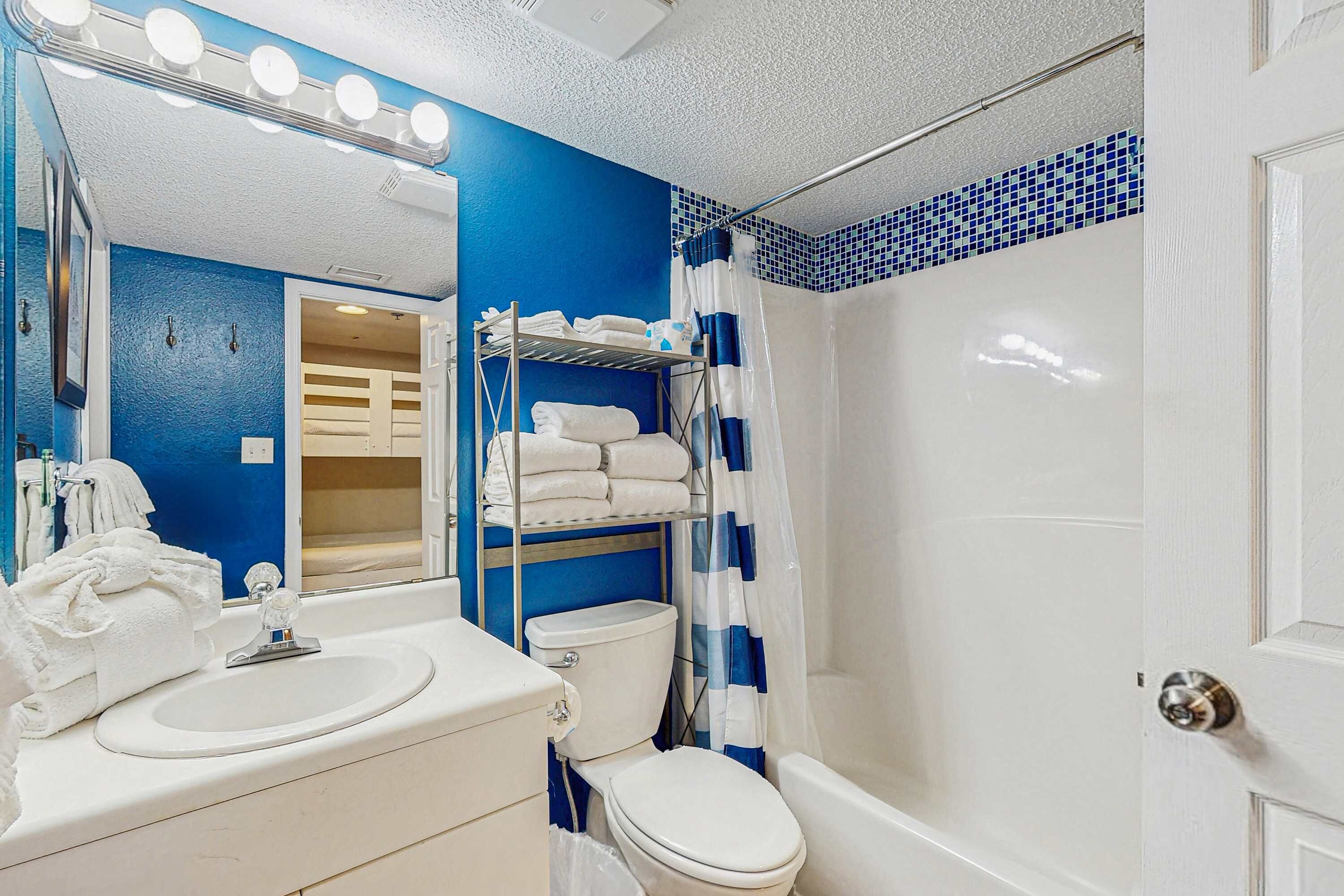 A bathroom with a blue wall, a white sink, a toilet, a shower with a blue and white curtain, and a mirror with lights.