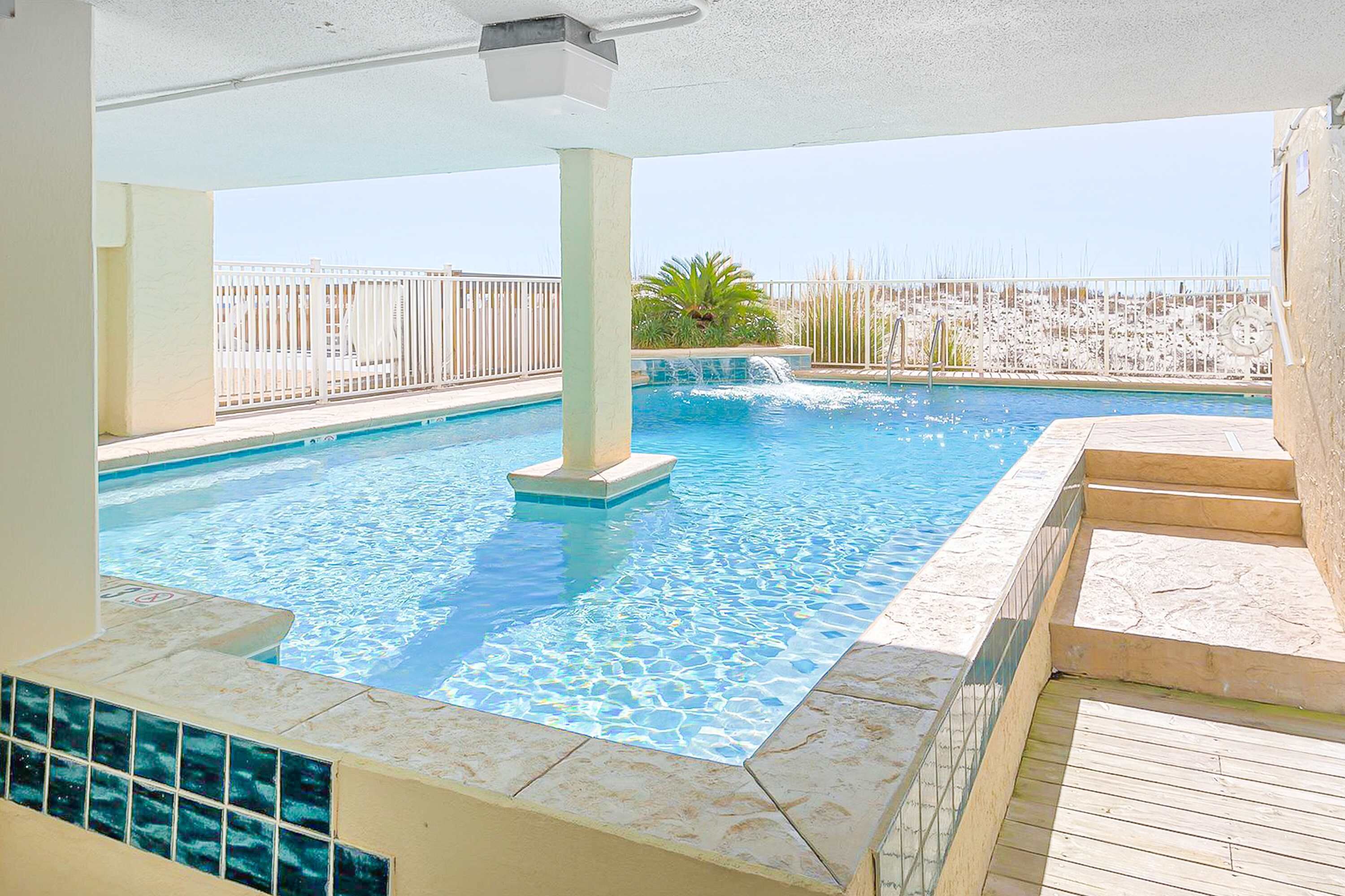 A swimming pool with clear blue water, surrounded by a white fence and a palm plant.