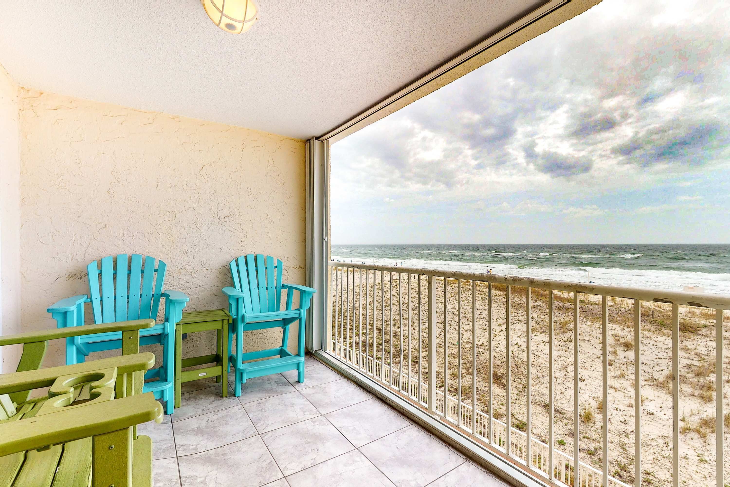 Varanda com duas cadeiras Adirondack verdes e uma mesa pequena, com vista para uma praia arenosa e o oceano.