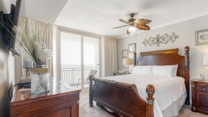 A bedroom with a wooden bed, a ceiling fan, a large window, and a desk with a vase. - Beachfront condo with pool, hot tub, balcony, fitness room & amazing corner view (Gulf Shores)