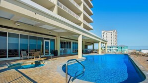 A swimming pool with a metal ladder, outdoor seating, and a view of a tall building in the background. - Beachfront condo with pool, hot tub, balcony, fitness room & amazing corner view (Gulf Shores)