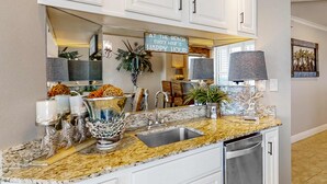 A kitchen with a granite countertop, white cabinets, a stainless steel sink, and a decorative sign. - Beachfront condo with pool, hot tub, balcony, fitness room & amazing corner view (Gulf Shores)