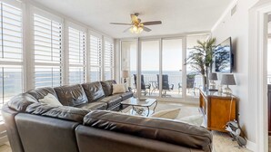 A spacious living room with a leather sofa, a ceiling fan, and a view of the ocean through large windows. - Beachfront condo with pool, hot tub, balcony, fitness room & amazing corner view (Gulf Shores)