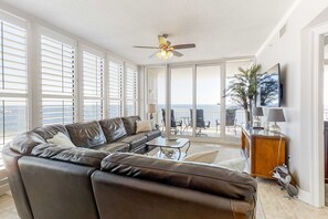 A spacious living room with a leather sofa, a ceiling fan, and a view of the ocean through large windows. - Beachfront condo with pool, hot tub, balcony, fitness room & amazing corner view (Gulf Shores)