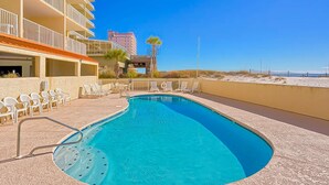 A swimming pool with a curved edge, surrounded by lounge chairs and a building with balconies. - Gulf-front 3rd-floor condo with outdoor pool, central AC, & balcony (Gulf Shores)