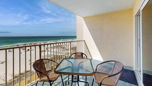 A balcony with a glass table and wicker chairs, overlooking a beach with umbrellas and people. - Gulf-front 3rd-floor condo with outdoor pool, central AC, & balcony (Gulf Shores)