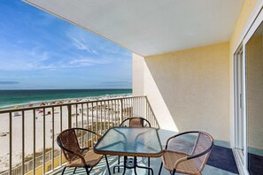 Ein Balkon mit Glastisch und Korbstühlen, der Blick auf einen Strand mit Sonnenschirmen und Menschen fällt.