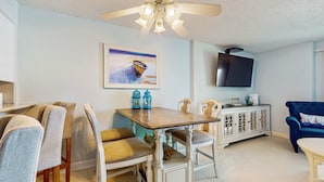 A dining area with a wooden table and chairs, a ceiling fan, a TV mounted on the wall, and a cabinet with decorative items. - Gulf-front 3rd-floor condo with outdoor pool, central AC, & balcony (Gulf Shores)