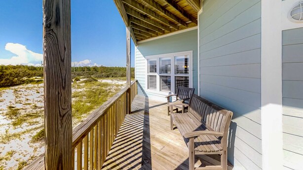 Eine Holzterrasse mit Blick auf den Strand, ein überdachter Bereich mit einer Bank und ein Fenster mit Rollläden.