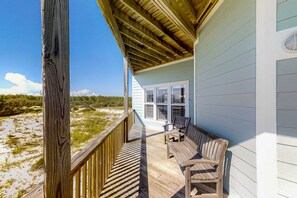 Terraza de madera con vista a la playa, zona cubierta con un banco y ventana con contraventanas.