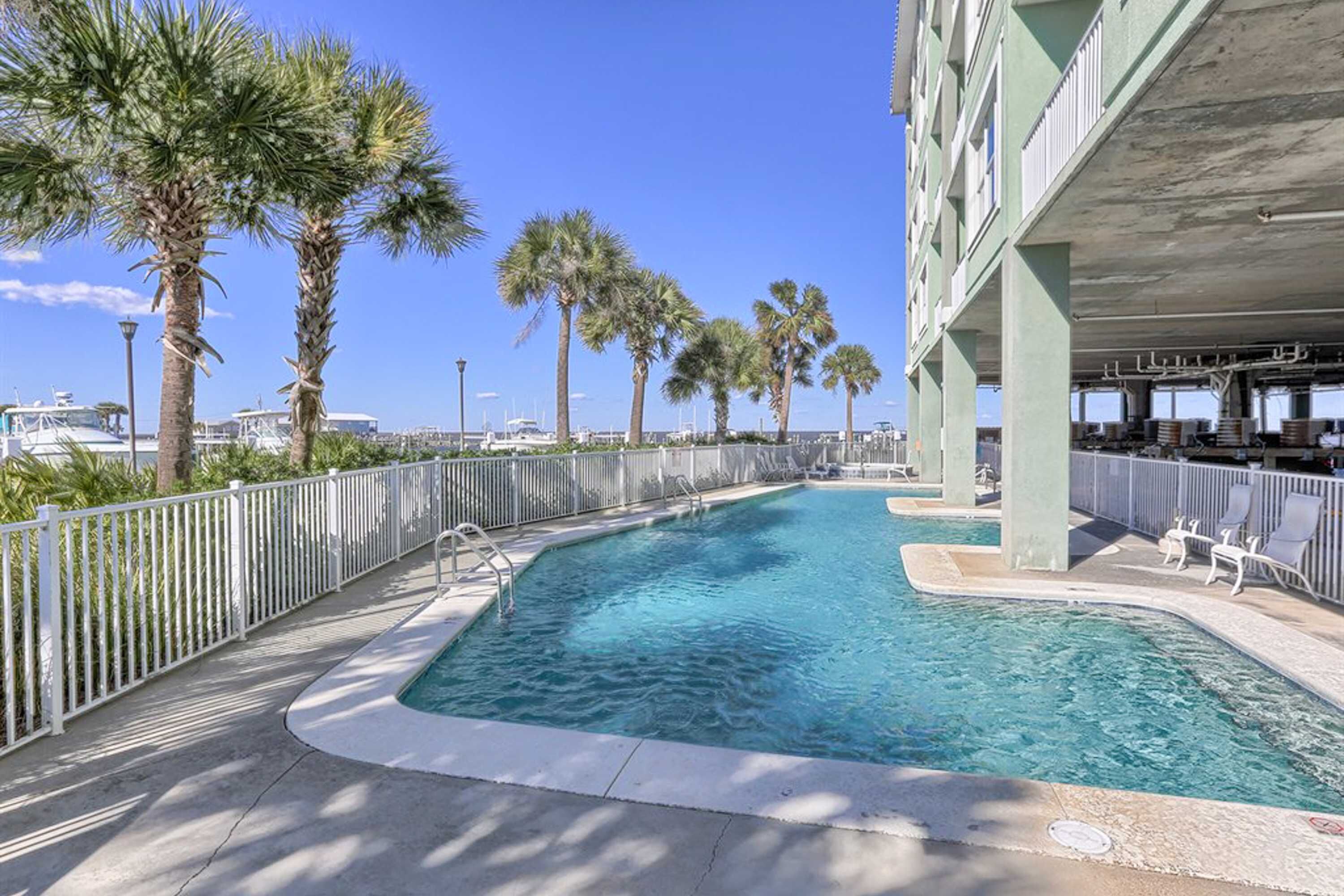 A swimming pool with a curved edge, surrounded by a white fence, palm trees, and a building with balconies.