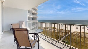 A balcony with a table and chairs, a white railing, and a view of the beach. - Gulf-Front Condo w Big Beach Views, Balcony, WiFi & Shared Pools, Hot Tubs & Gym (Gulf Shores)