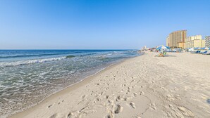 Une plage parsemée d’empreintes, un ciel dégagé et des bâtiments en arrière-plan.