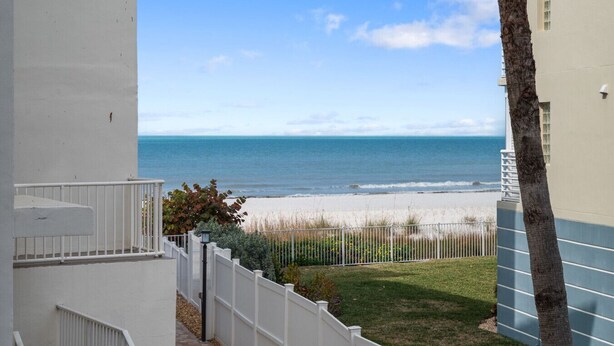 Uitzicht op een strand vanaf een balkon met een witte railing, een palmboom en een hek in de voorgrond.