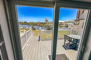 A wooden deck with a white railing, a table and chairs, and a view of a river and buildings in the distance. - Waterview getaway with Private Pier, Balcony and Kayaks (Gulf Shores)