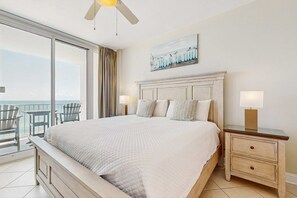 Une chambre d’hôtel avec un grand lit, deux tables de chevet, un ventilateur de plafond, un balcon avec des chaises et un tableau au mur.