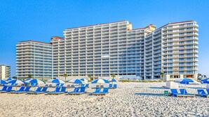 Un grand hôtel à étages multiples et doté de balcons, une plage de sable fin avec des chaises longues bleues et des parasols, et un ciel bleu azur.