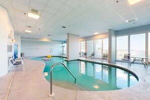 An indoor swimming pool with a curved design, surrounded by lounge chairs and a view of the city. - Bright beachfront condo w private balcony, zero-entry pool, gym, & hot tub (Gulf Shores)