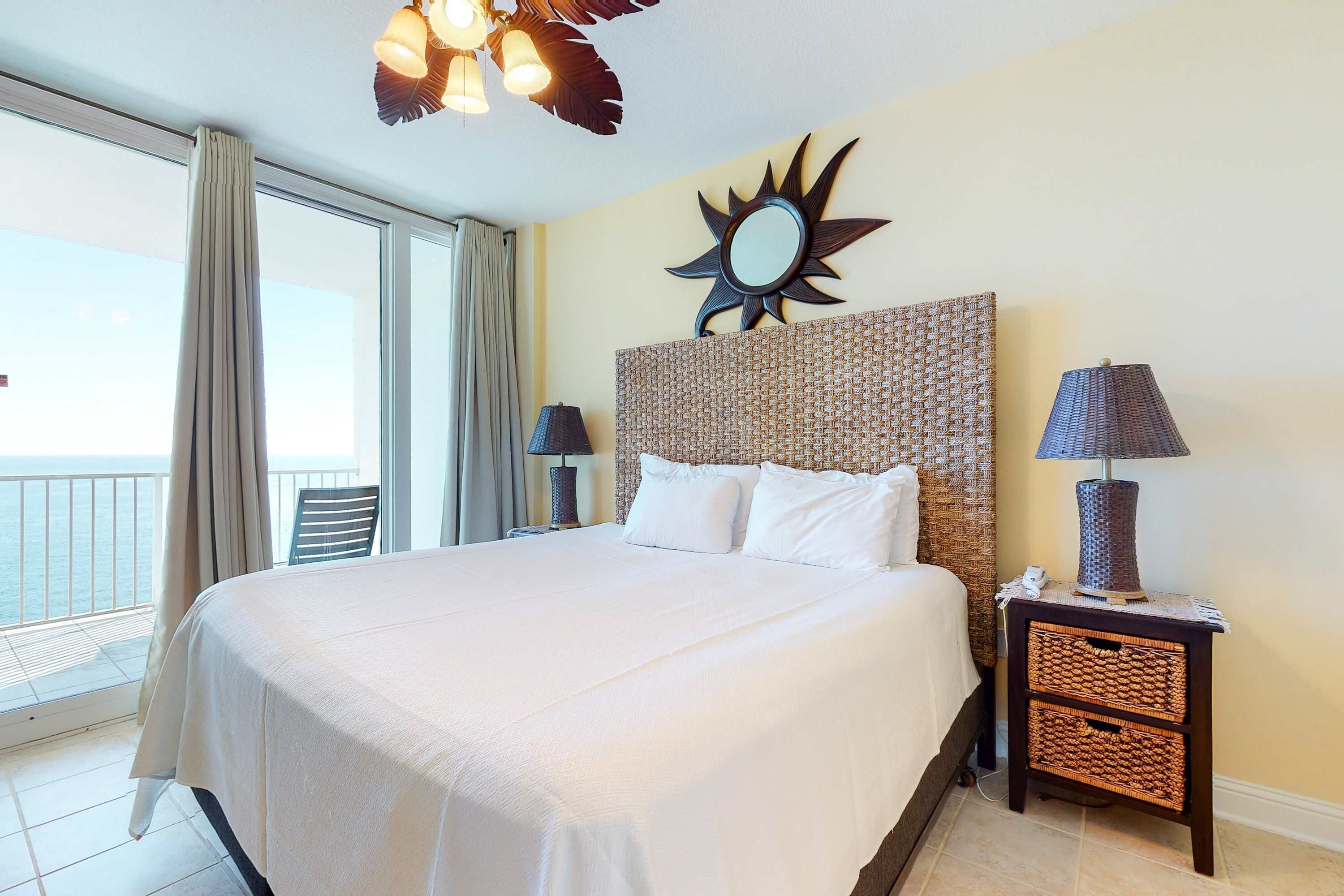 Habitación de hotel con una cama grande, un espejo con forma de sol en la cabecera, una mesita de noche con lámpara y vista al mar a través de la ventana.