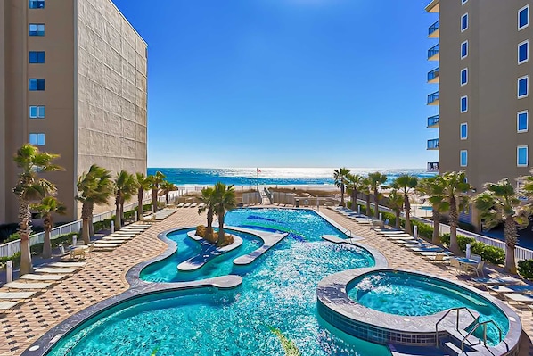 A pool area with multiple swimming pools, palm trees, and a view of the beach. - Gorgeous Gulf view condo with indoor,outdoor pools, hot tubs, gym, & balconies (Gulf Shores)