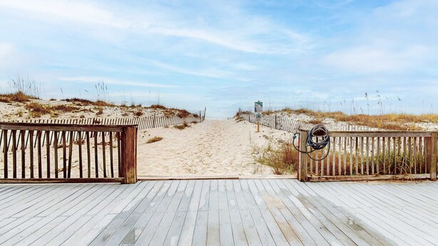 A wooden boardwalk leading to a sandy beach with a fence on one side and a signpost on the other. - Beachfront condo with outdoor pool, beach views, washer/dryer, beach access (Gulf Shores)