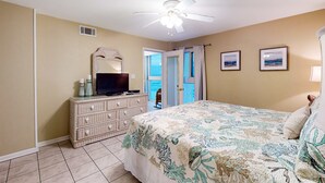 Une chambre à coucher comprenant un lit, une commode, une télévision, un ventilateur de plafond et une porte donnant sur un balcon.
