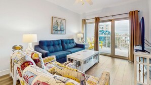 A living room with a blue sofa, a white coffee table, a patterned armchair, a painting of sailboats, and a balcony with a view of a building and a blue structure. - Gulf view condo with 2 pools, lazy river, hot tub, sauna, gym & beach access (Gulf Shores)
