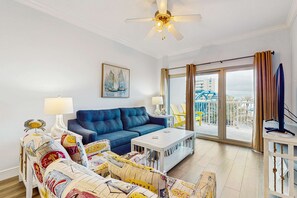 A living room with a blue sofa, a white coffee table, a patterned armchair, a painting of sailboats, and a balcony with a view of a building and a blue structure. - Gulf view condo with 2 pools, lazy river, hot tub, sauna, gym & beach access (Gulf Shores)