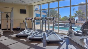 A gym with treadmills, an elliptical, and a rowing machine, large windows, and a view of a pool and buildings. - Gulf view condo with 2 pools, lazy river, hot tub, sauna, gym & beach access (Gulf Shores)