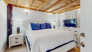 Quarto com teto de madeira, cama com roupa de cama branca e azul, televisão na parede e mesa de cabeceira com abajur.