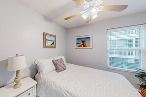 Quarto com cama, mesa de cabeceira, abajur, ventilador de teto, duas fotos emolduradas e janela com persianas.