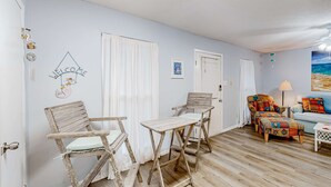 Una habitación con piso de madera, dos sillas de madera, una mesita, un sillón colorido, un sofá y un cuadro de una playa.