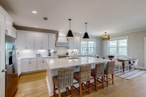A modern kitchen with white cabinetry, a large island with bar stools, pendant lighting, and a view of the outside. - Sprawling retreat with an enclosed pool, large balcony, outdoor shower, & W&D (Gulf Shores)