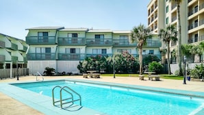 A swimming pool with a metal ladder, surrounded by a fence, with a multi-story building in the background. - Renovated condo by the beach with pool access & private balconies (Gulf Shores)