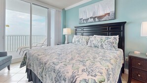 Een slaapkamer met een bed, nachtkastje, lamp, schilderij, een balkon met stoelen en uitzicht op zee.