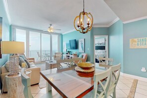 A spacious living area with a dining table, chairs, a chandelier, and a view of the outdoors. - Oceanfront condo with sweeping views, washer/dryer, central AC, & access to pool (Gulf Shores)