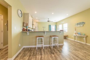 A kitchen with wooden flooring, white cabinets, two stools, a ceiling fan, and a clock on the wall. - Pristine beach home with gulf view, covered patio, and AC (Gulf Shores)