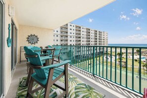 Ein Balkon mit türkisfarbenen Stühlen und einem dekorativen Wandelement, mit Blick auf einen Strand und ein mehrstöckiges Gebäude.