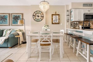 A modern kitchen with white cabinetry, a dining area with a white table and chairs, and a living room with a teal sofa and artwork on the wall. - Beachfront condo with Gulf views, pools, hot tubs, tennis courts, gym, AC, & W&D (Gulf Shores)