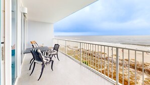 Een balkon met een tafel en stoelen, met uitzicht op een strand met een hek en een zeezicht.