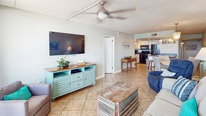 A living room with a TV, a dining area, and a kitchen. - Gulf front condo with stunning Gulf views, outdoor pool & beach access (Gulf Shores)